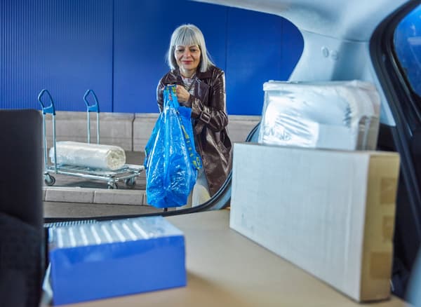 Naeratav inimene tõstab oste täis sinist FRAKTA kotti auto pagasiruumi. Tema taga on ostukäru peal rullikeeratud madrats.