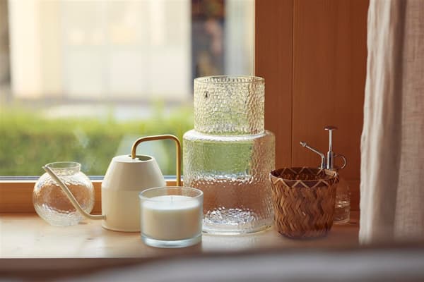 Nahaufnahme einer Fensterbank mit Vasen, einer kleinen, eleganzen Giesskanne, einer Duftkerze und einem Wasserzerstäuber