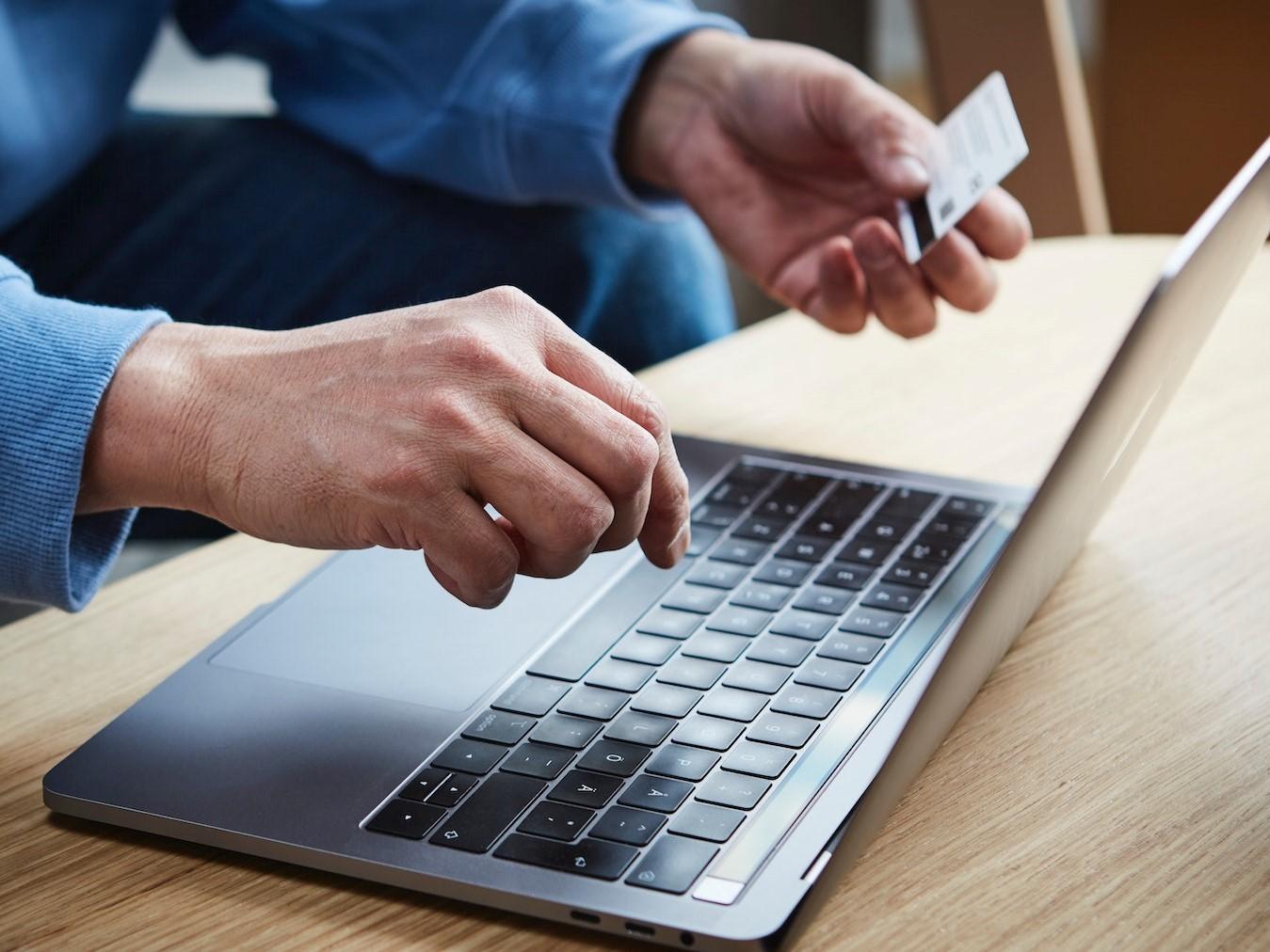 Nahaufnahme eines aufgeklappten Laptops an dem eine Person mit einer Kreditkarte in der Hand sitzt