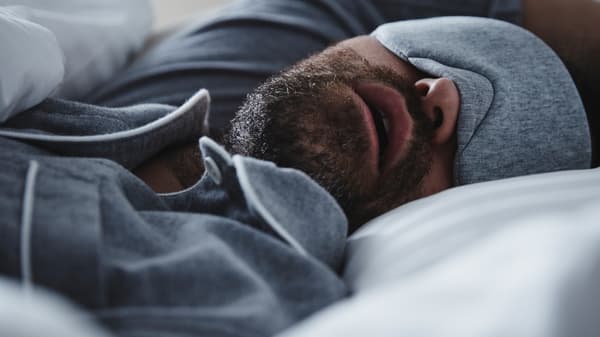 גבר בפיג'מה אפורה וכיסוי עיניים תואם ישן בפה פתוח במיטה עם מצעי NATTJASMIN לחדר השינה.