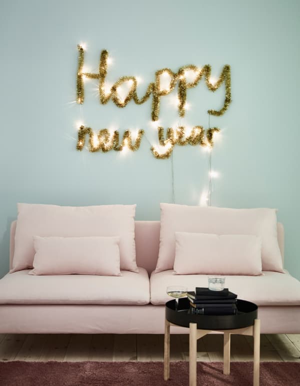 New Year wall decoration made using SÄRDAL battery-operated LED light chains and a gold garland, above a pink sofa.