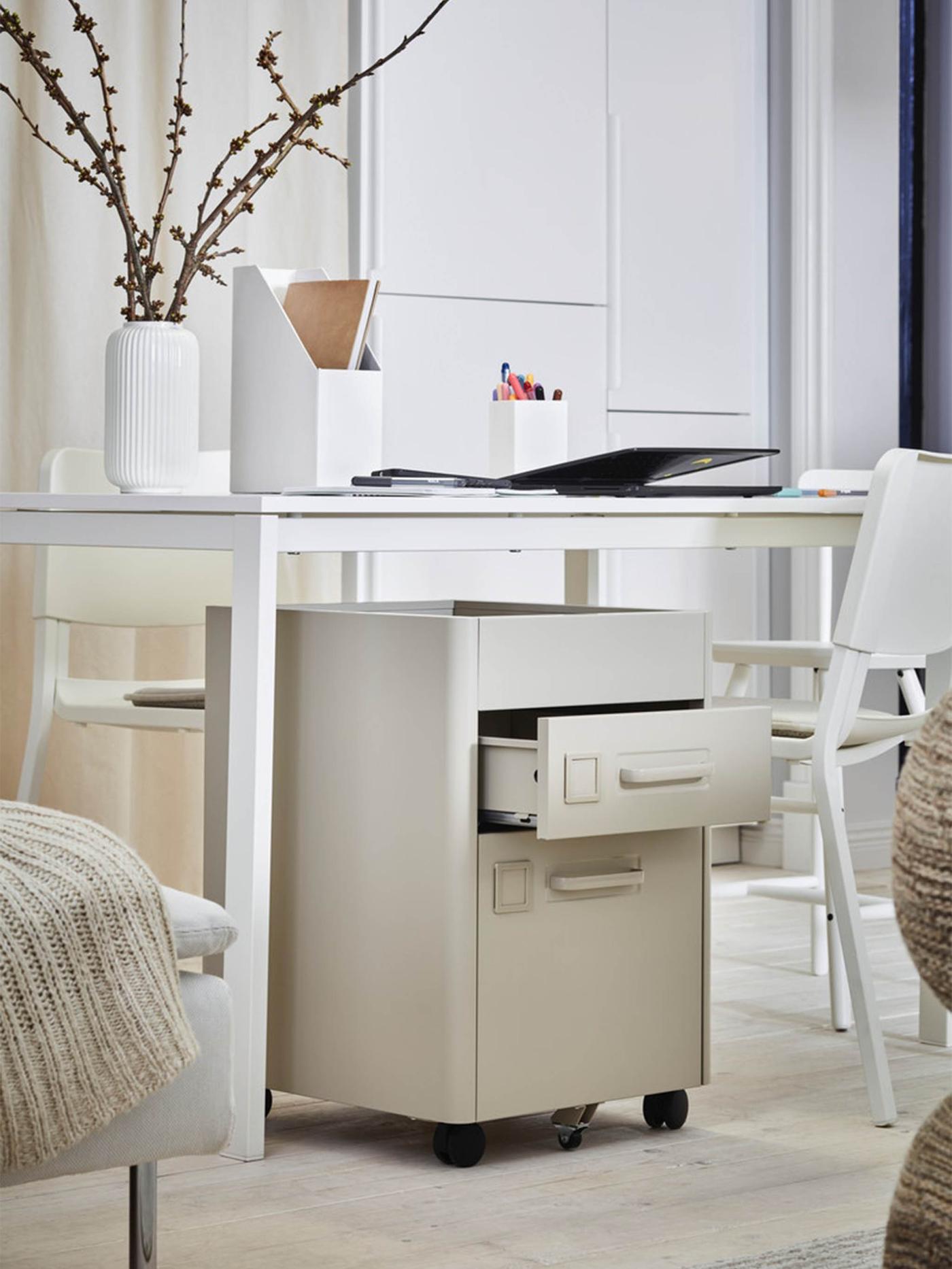 Next to a bed stands a white table with work accessories scattered on it and an IDÅSEN drawer unit placed underneath.