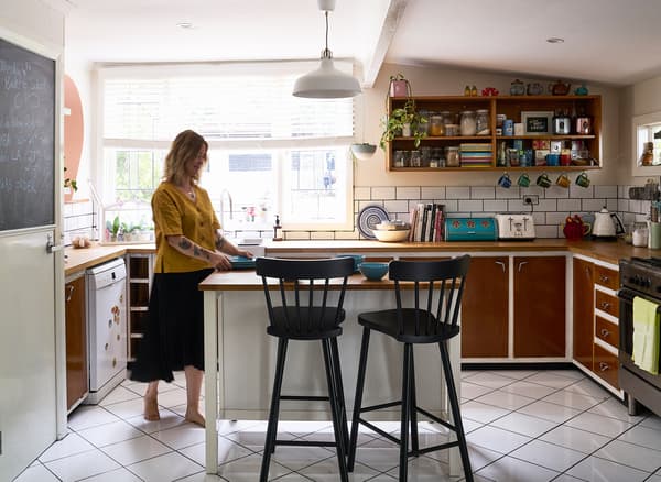 Nici standing at an island with two black bar stools, in a kitchen with brown cupboards, shelves and a large window.