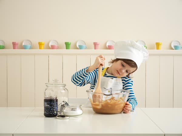 Niña  con una polera a rayas azules y blancas. Usando un gorro de chef y un delantal blanco mientras revuelve una masa dentro de un bowl de vidrio IKEA.