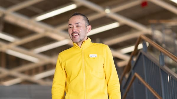 黄色のユニフォームを着たスタッフが立つ職場シーン。背景に木材や構造材が見える広々とした空間。