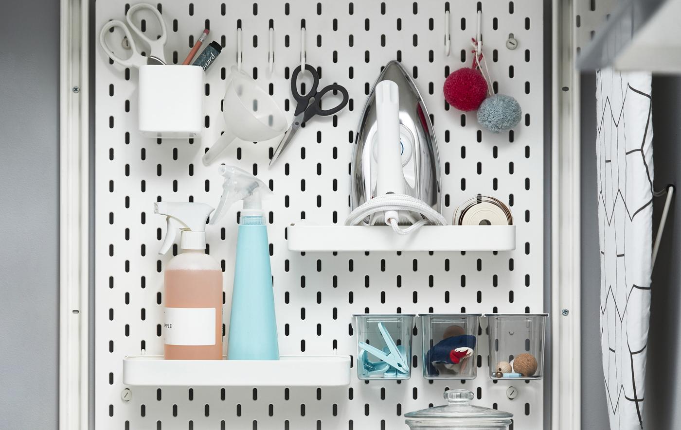 Objets en rapport avec la buanderie rangés sur des tablettes, dans des pots et à des crochets fixés sur un panneau perforé blanc.
