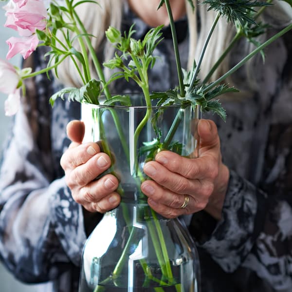 OMTÄNKSAM vase, with beautifully arranged stems, held by an elderly woman. The shape makes the vase easy to lift and carry.