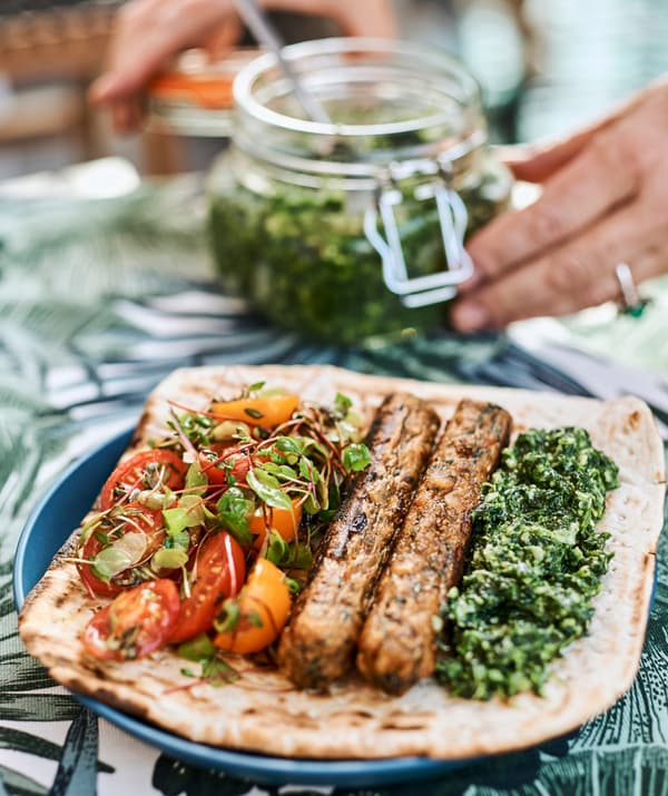 On a blue plate, flatbread with pesto, sausages and tomato salad, hands close a glass jar of pesto.