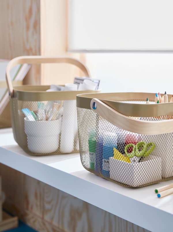 On a sunlit windowsill stand two light olive-green RISATORP baskets, holding various drawing and watercolour accessories.