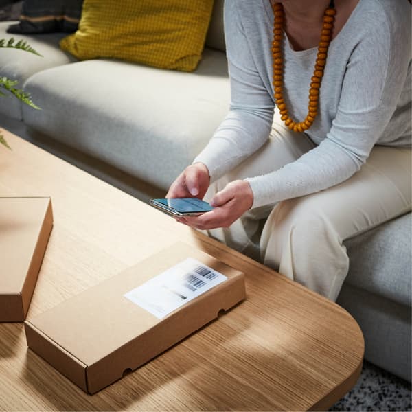 Op een lichte houten IKEA salontafel scant een persoon in een grijze top en crèmekleurige broek een pakketbarcode met hun smartphone, terwijl comfortabel zittend op een moderne grijze IKEA bank met een groene plant ernaast.