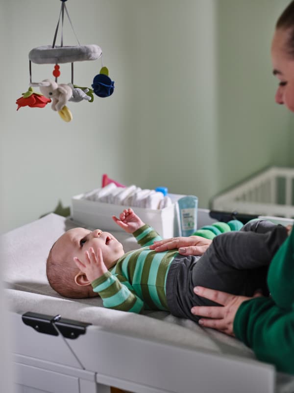 Op een SUNDVIK commode verzorgt een volwassene een aangeklede baby die kijkt naar de veelkleurige GULLIGAST mobiel die boven hem hangt.