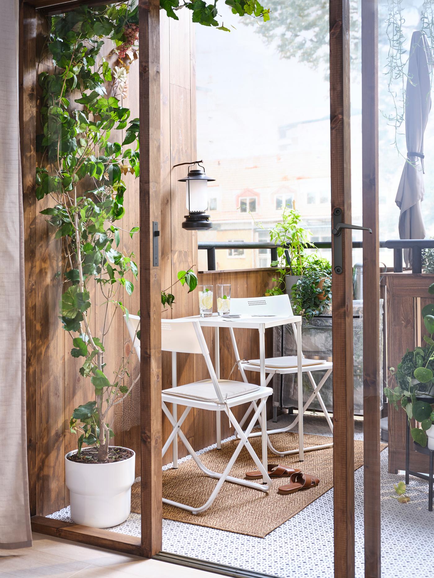 Open doors leading to a leafy balcony which has a white table and two chairs on it, with a lantern on the wall above.