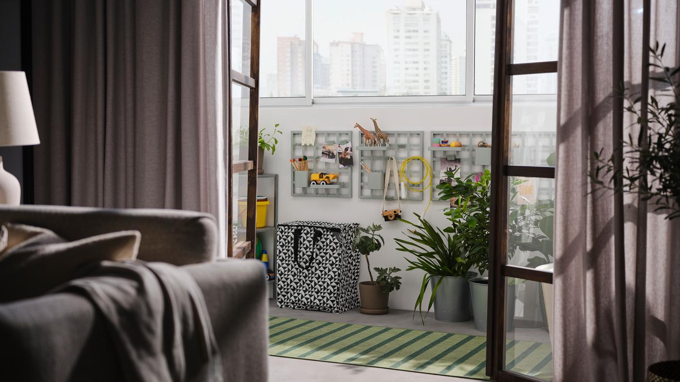 Open doors onto a balcony with grey-green SNICKRA storage boards on a wall, a black/white bag, large potted plants and a rug.