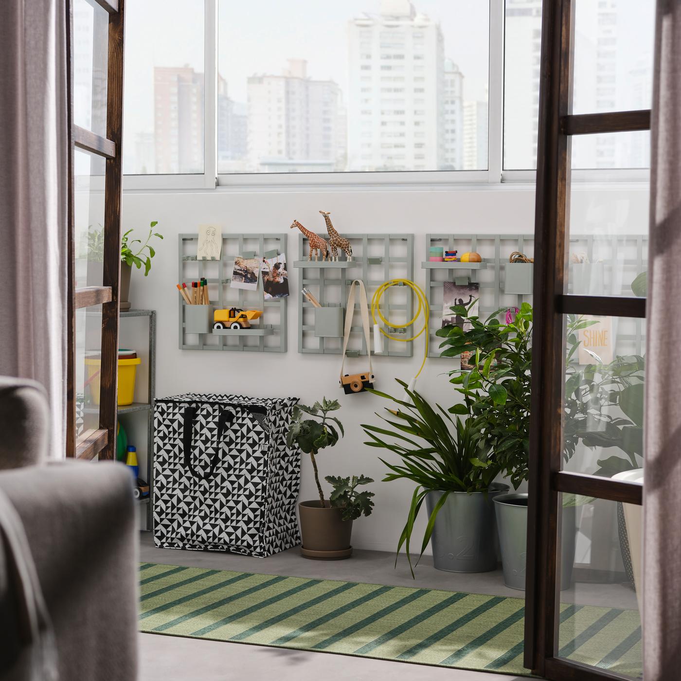 Open doors onto a balcony with grey-green SNICKRA storage boards on a wall, a black/white bag, large potted plants and a rug.