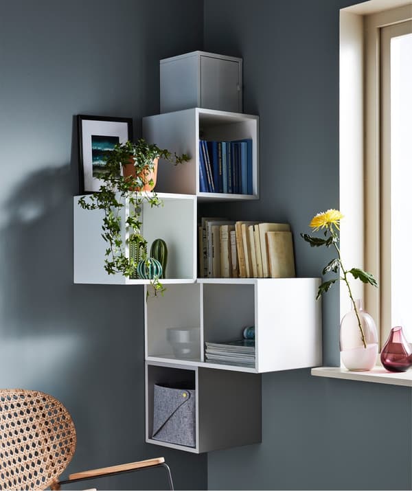 Open wall storage units, in a corner of a grey room, decorated with books, plants and picture frames.