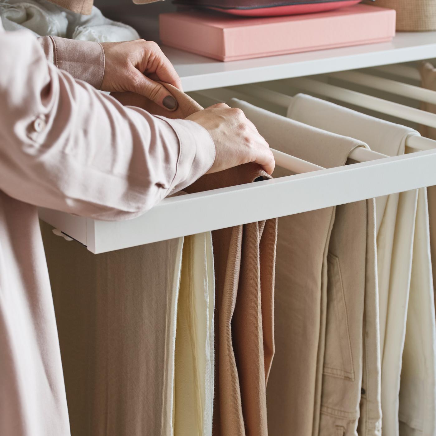 Osoba u roze košulji podešava bež pantalone na jednoj od nekoliko belih KOMPLEMENT izvlačnih vešalica za pantalone.