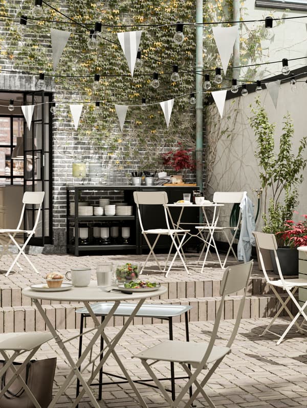 Outdoor dining in the courtyard with white folding tables and chairs, string lights and pennants above, and a serving area against an ivy-covered wall with plants around it.