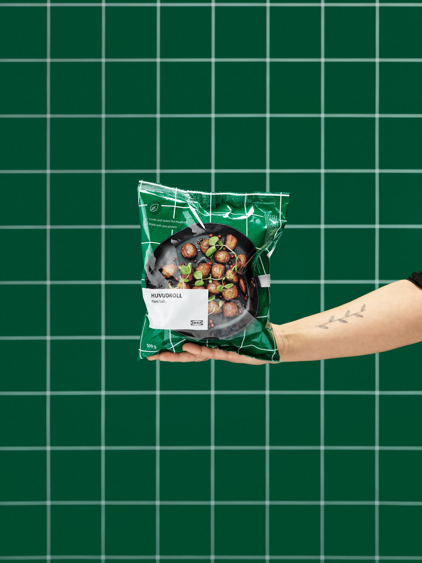 Outstretched hand holding an unopened package of HUVUDROLL plant balls. Green, tiled wall in the background.