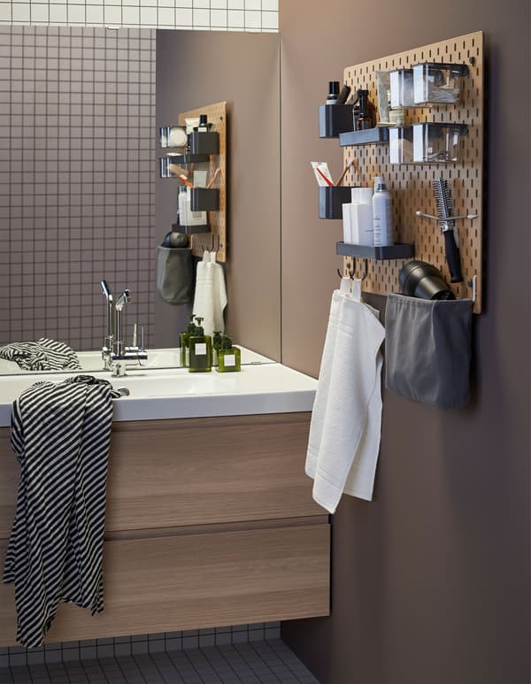 Panneau perforé en bois SKÅDIS installé à côté d’un lavabo, dans une salle de bains, et garni d’accessoires pour les brosses à dents et autres articles de toilette.