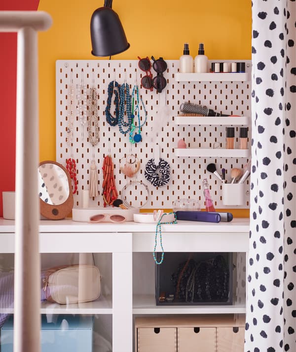 Panneau perforé fixé au mur. Sur ses crochets et tablettes sont disposés des colliers, bracelets, lunettes de soleil et cosmétiques.