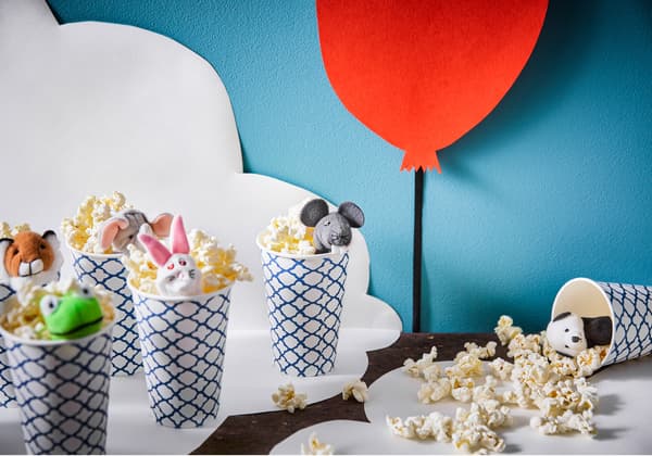 Paper cups are filled with popcorn and topped with a finger puppet. One cup is tipped over with popcorn spilling out.