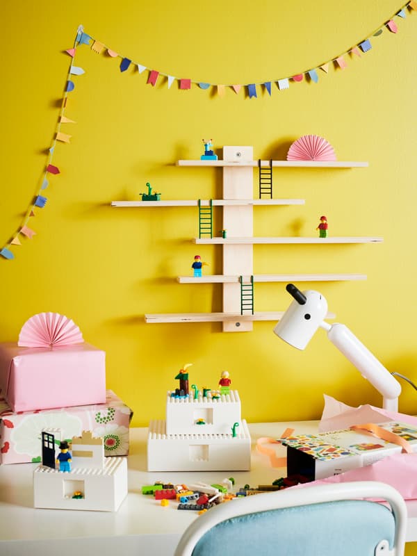 Pared amarilla con repisas blancas con un soporte de madera en una pieza infantil. Sobre ella hay juguetes y está sobre un escritorio blanco infantil con legos.