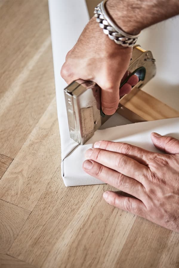 Parquet sur lequel un homme agrafe un morceau de toile blanche sur le coin d’un cadre en bois.