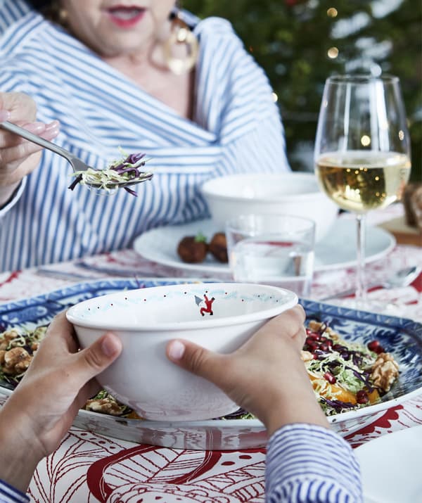 Partie d’une table de fête, avec assiettes et verres remplis. Une personne âgée dépose de la nourriture dans un bol tenu par un enfant.