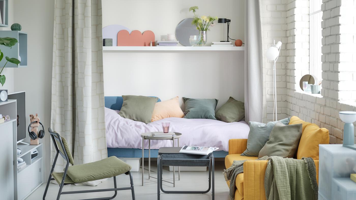 Pastelkleurige woonkamer waar je elk uur van de dag terechtkan, met BLÅKULLEN bed in middenblauw, gele slaapbank en olijfgroene fauteuil.