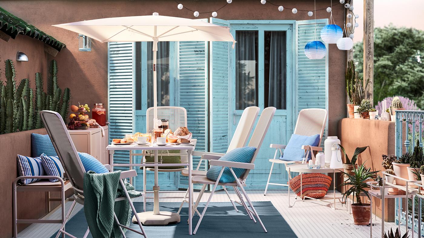 Patio yang disinari cahaya matahari dengan dinding terracotta, pintu berwarna biru, meja dengan payung berwarna putih, kerusi luar berwarna putih dan kusyen berwarna biru.
