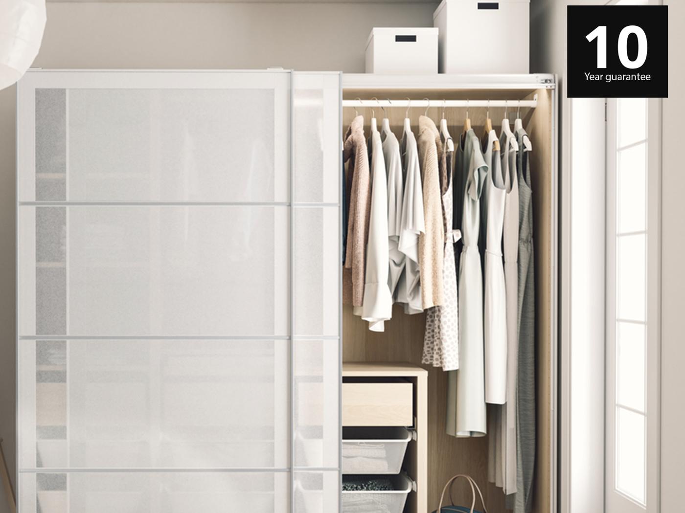 PAX wardrobe in bedroom with SVARTISDAL sliding doors. One side of the wardrobe is open with clothes hanging on BUMERANG hangers and there are 3 mesh drawers and a bag at the bottom of the wardrobe.