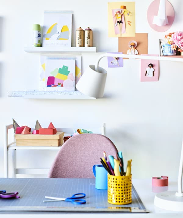 Pen pots on a desk with a white desk lamp, pink chair, and artwork on picture ledges on the wall behind.