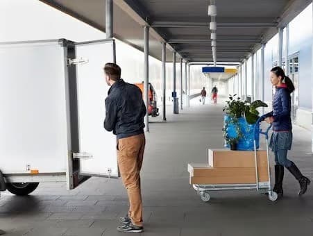 Person unloading from transport cart while another person drives a cart with boxes in a covered aisle.