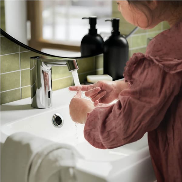 Person washing hands under running water at a modern bathroom sink. Soap dispensers and green tiles create a fresh, clean setting.