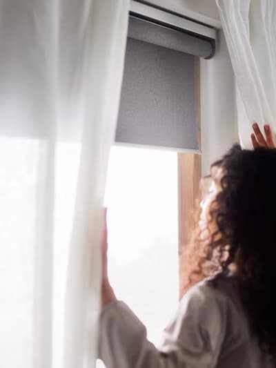 Person with curly hair opening sheer white curtains next to a window with a gray roller blind, sunlight streaming into the room.