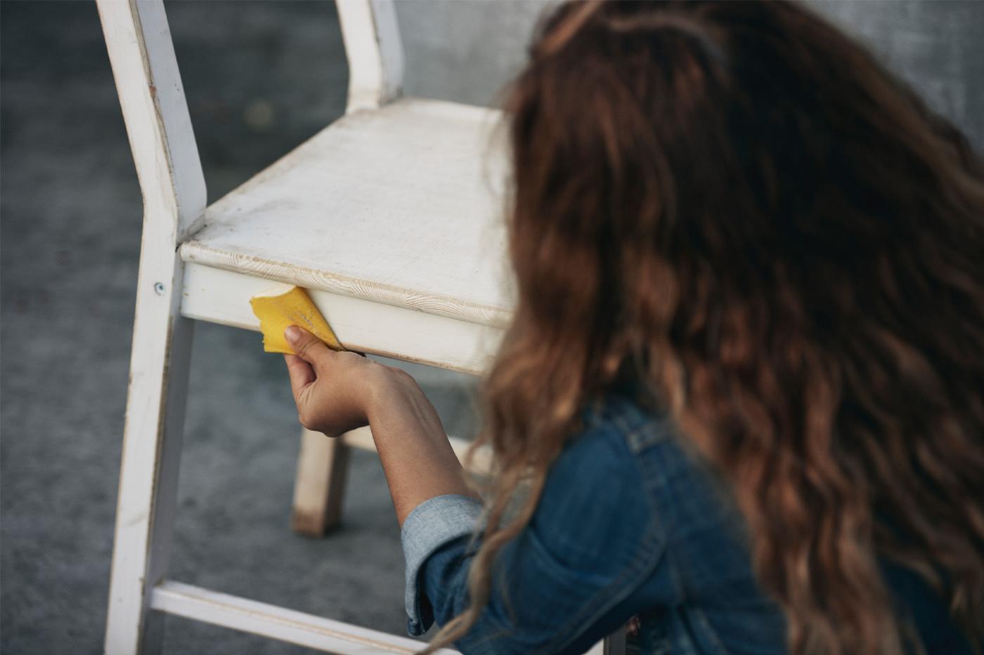 Person with long brown hair using sandpaper to sand a white-painted wooden chair standing on a gray floor.