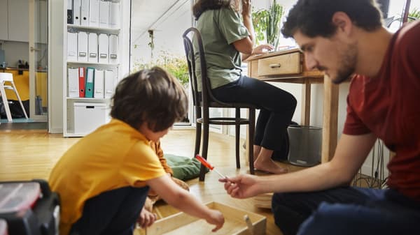 Person works at a desk while another person and child assemble furniture on the floor in a bright home office.