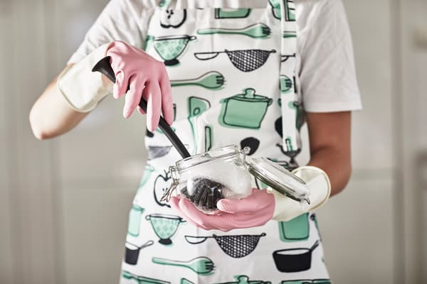 Persona con un delantal y guantes limpiando un frasco de vidrio con un utensilio de limpieza IKEA.