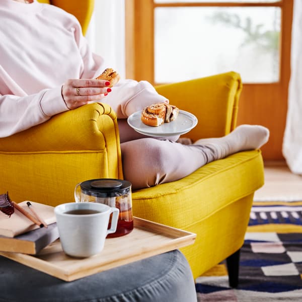 Persona sentaba en un sillón tapizado STRANDMON amarillo sosteniendo un plato con un rollito de canela IKEA.