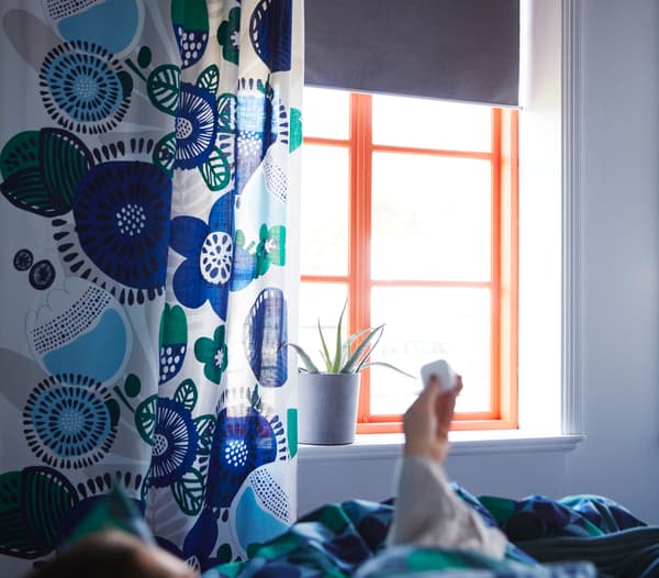Personne ouvrant des stores connectés FYRTUR depuis son lit à l'aide d'une télécommande.