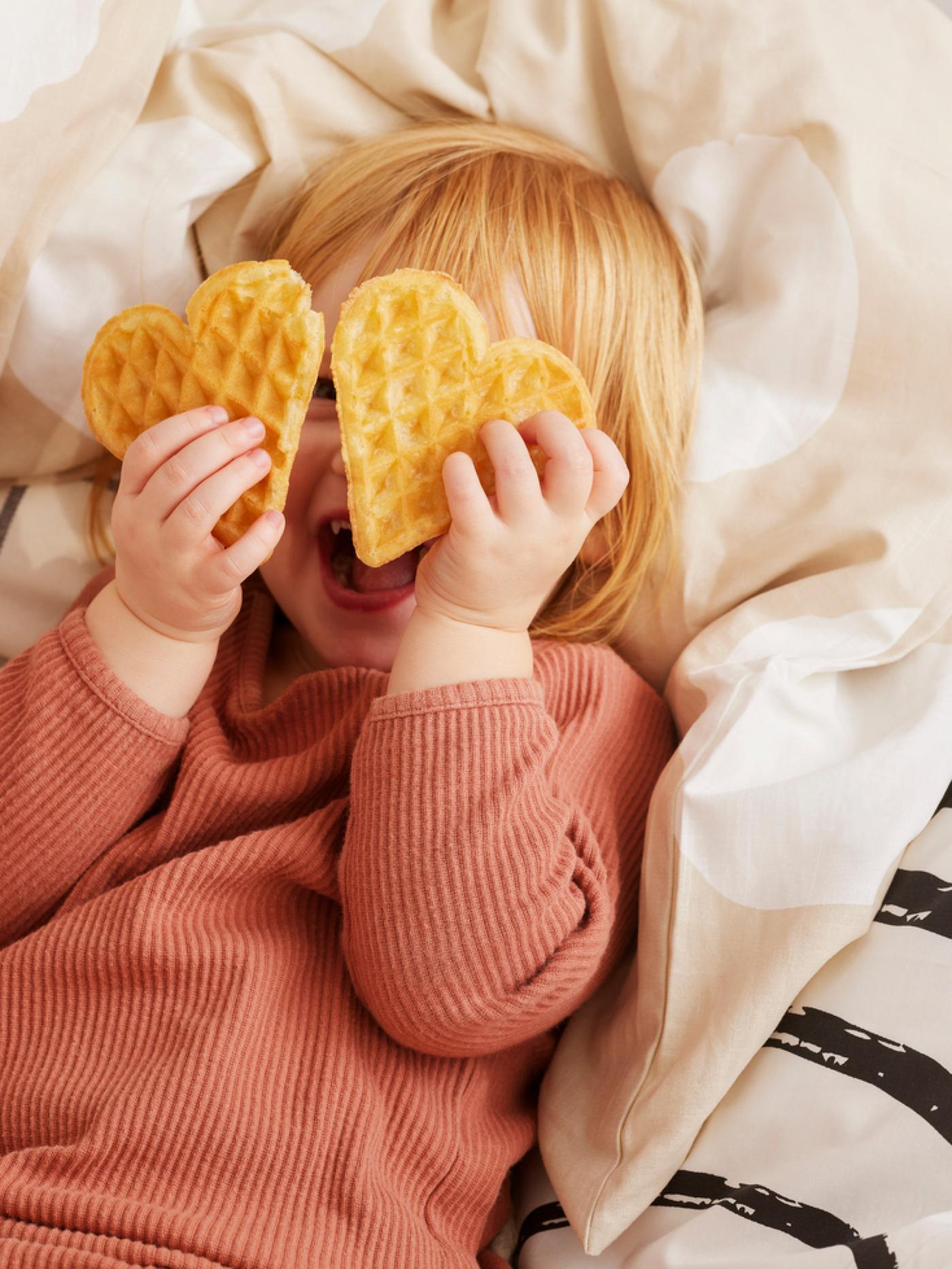 Petite fille sur un lit. Deux gaufres en forme de cœur devant son visage souriant.