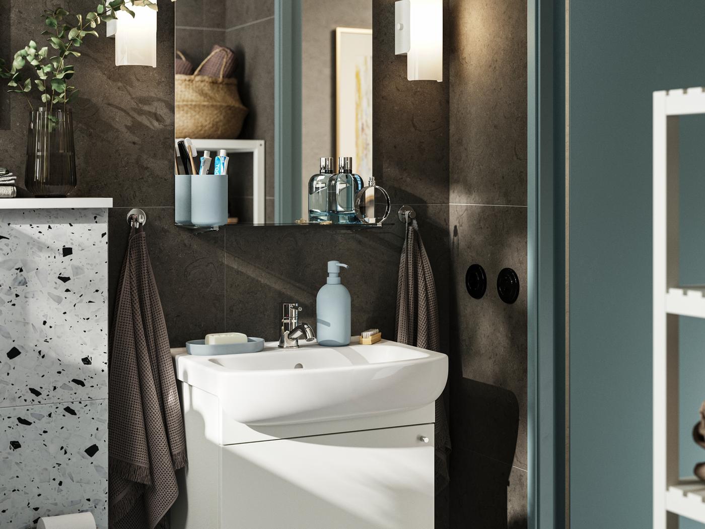Petite salle de bains aux murs brun foncé et aux meubles blancs. Armoire NYSJÖN à une porte contenant un rangement sous l’évier.