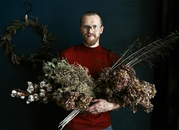 Photo de Łukasz debout devant une couronne, avec des bouquets de fleurs séchées dans les bras.