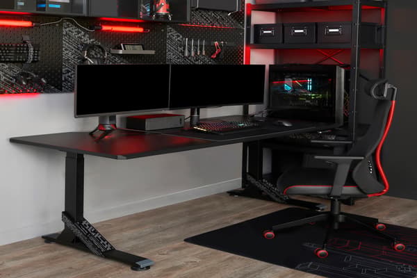 Picture of a desk with two screens and a red and black gaming chair in front of it.