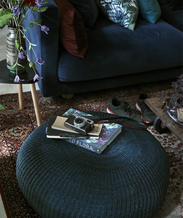 Picture of camera and books on a large round blue pouffe, with blue sofa in background.