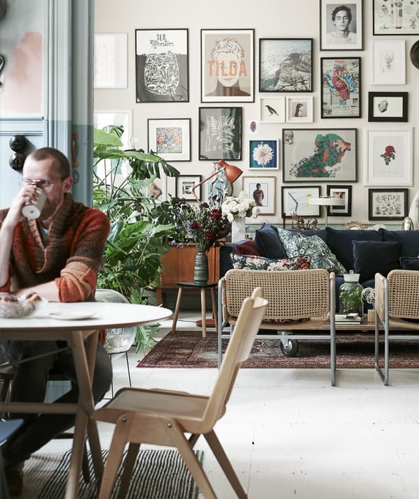 Picture of Łukasz sitting at a table drinking from a mug with view of the living room and wall of pictures in the background.
