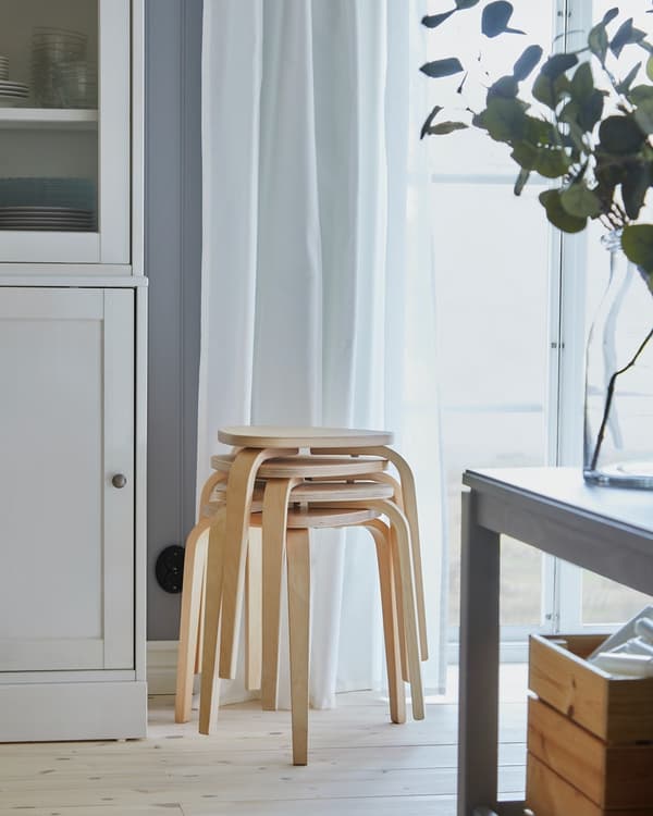 Pisos de madera montados uno sobre otros de la familia KYRRE de IKEA. Están puestos junto a la ventana, con una cortina blanca y un mueble blanco con puerta.