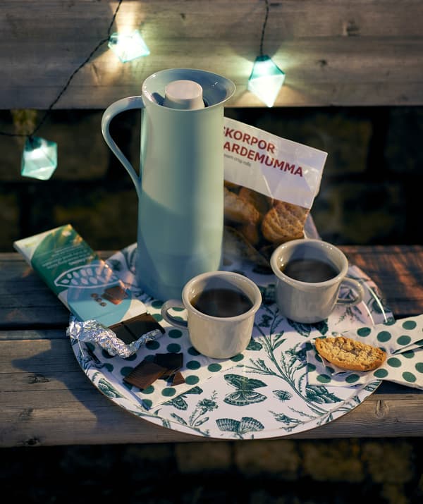 Placed on a night-time park bench, a MUSTIGHET tray holds a BEHÖVD vacuum flask, coffee mugs, crisp rolls and chocolate.