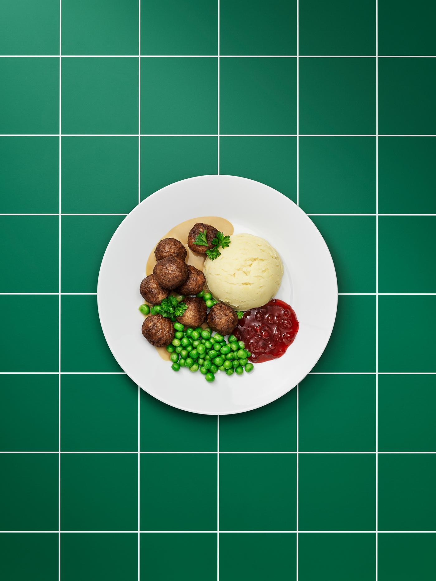 Plain white plate of plant-based meatballs, brown sauce, mashed potatoes, lingonberry jam, green peas and a sprig of parsley.