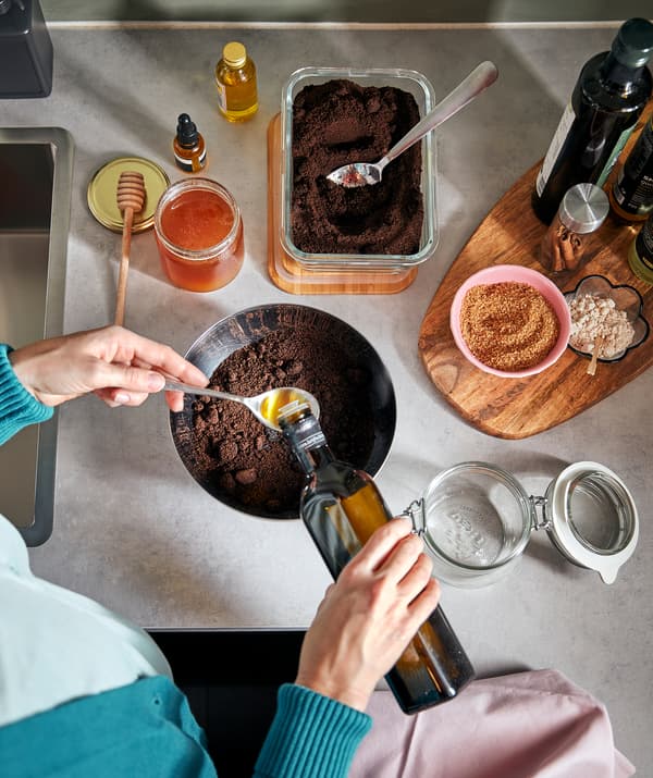 Plan de travail de cuisine avec une personne préparant un mélange en versant de l’huile au-dessus d’un bol rempli de café. Un bocal KORKEN vide se trouve sur le côté.